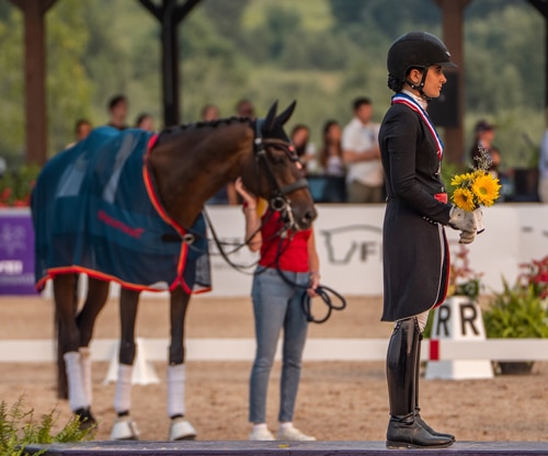 A woman standing on a podium with a bay horse in the background.