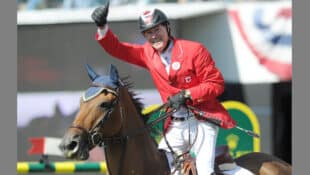 A man in a red jacket on a bay horse, waving to the crowd.