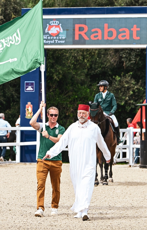 A man in a red fez and white robe leading a man with a flag into an arena.