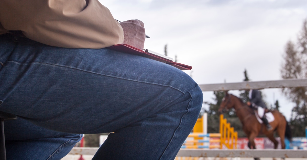 A man with a clipboard on his knee, judging a horse show class.