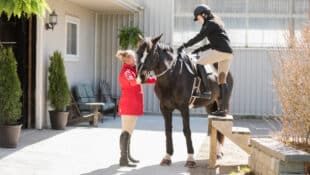 A woman holding a horse at a stable while another woman mounts the horse.