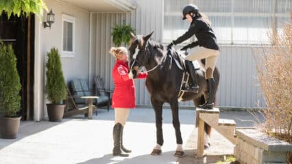 A woman holding a horse at a stable while another woman mounts the horse.