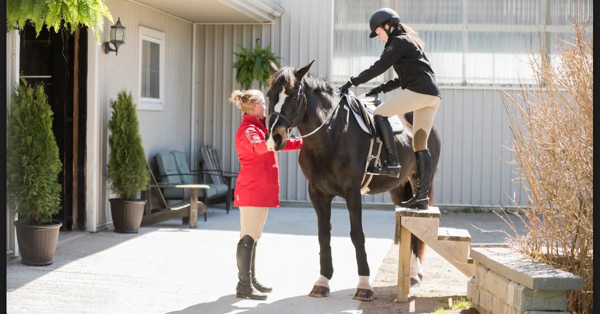 A woman holding a horse at a stable while another woman mounts the horse.