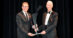 A man handing a trophy to another man in a tuxedo on a stage.