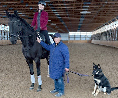 A womn standing in an arena with a mounted rider and a dog.