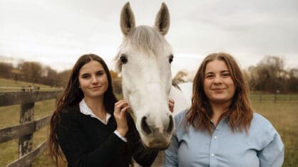 Two young women standing with a grey horse.