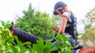 A teenage girl on a horse, looking at her cell phone.