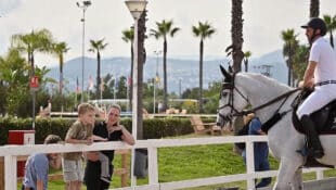 A blonde woman and 3 children watching a man on a horse.