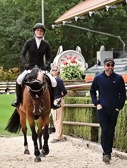 A woman on a horse with a man walking beside at the in-gate of a horse show.