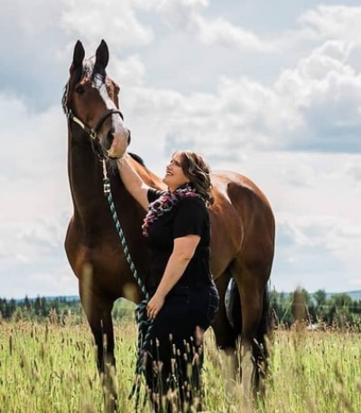 A woman standing i a field with a bay horse.