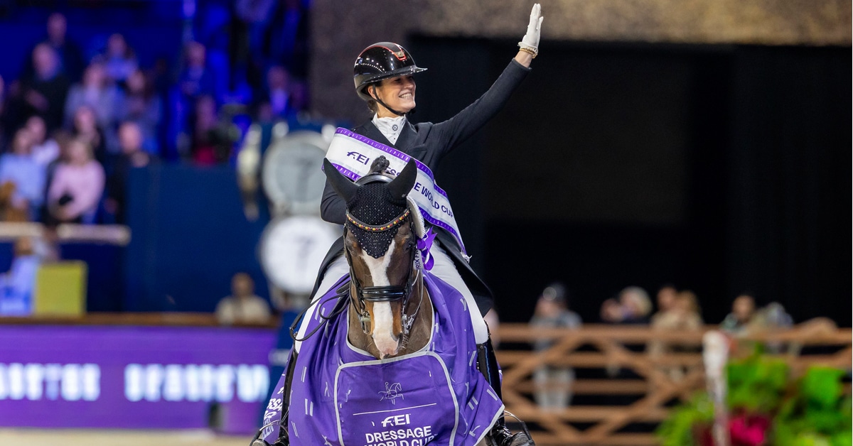 A woman on a bay dressage horse, smiling and waving to the crowd.