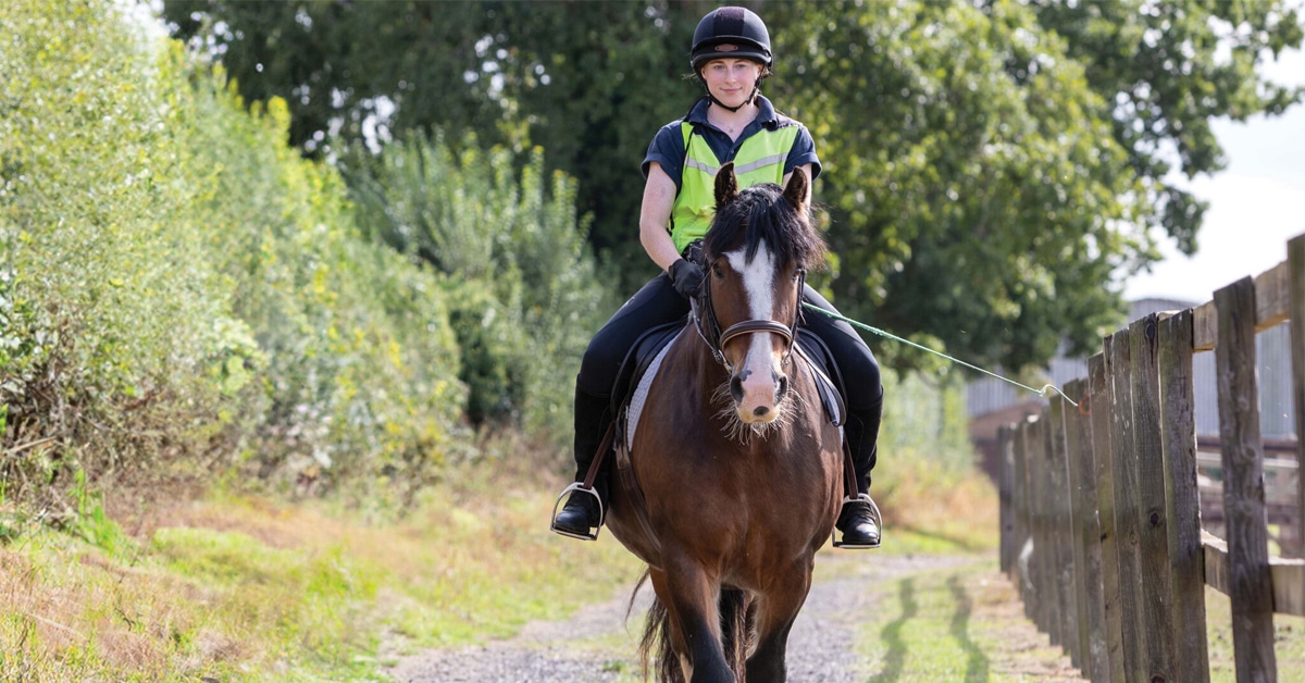 A woman carrying a whip, riding a bay horse along a road.