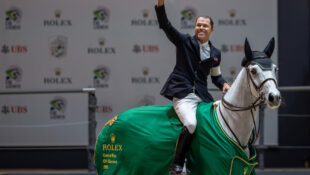 A man waving from the back of a grey mare that is wearing a Rolex cooler.