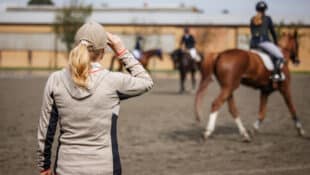 A female trainer watching horses and riders in a warmup ring.