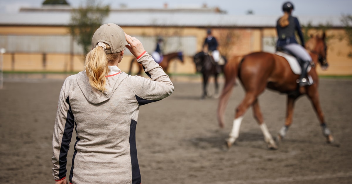 A female trainer watching horses and riders in a warmup ring.