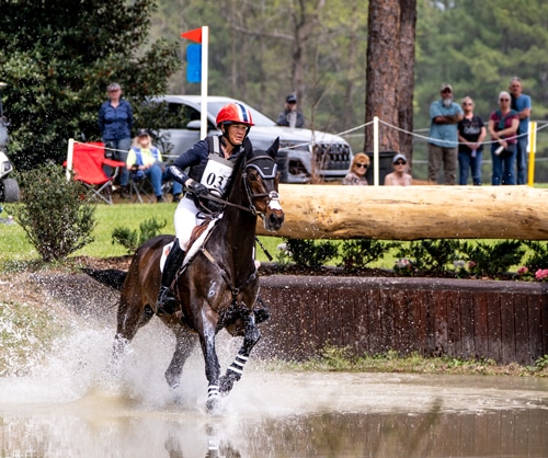 A woman galloping a dark bay horse trough a water obstacle.