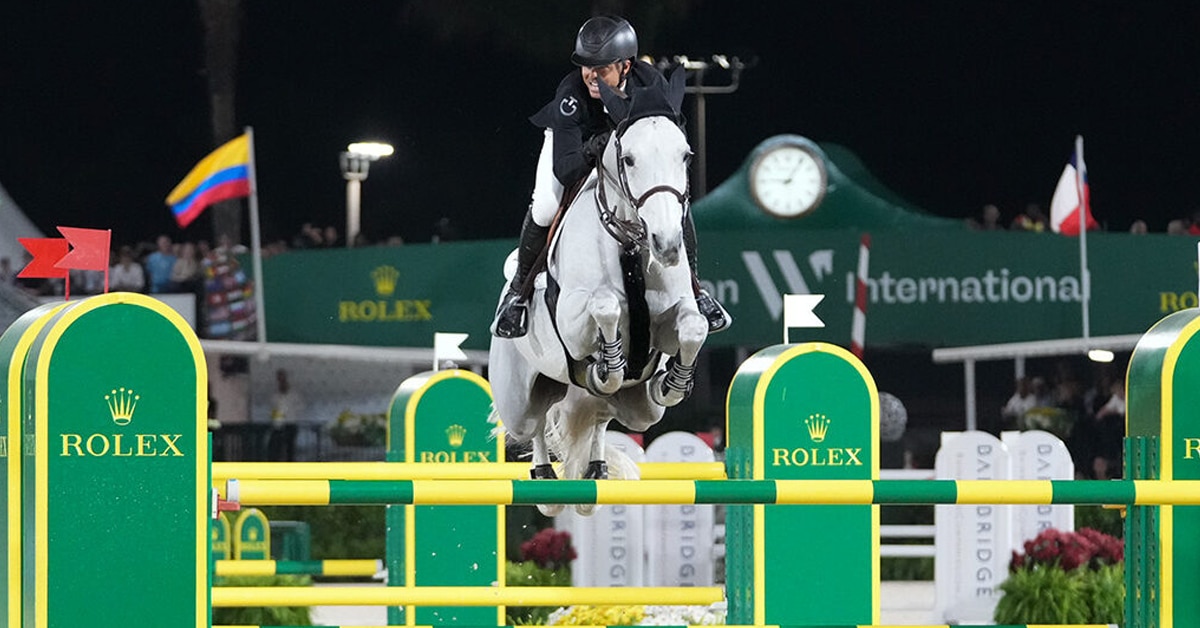 A man jumping a grey mare over a Rolex fence during a night class at WEF.