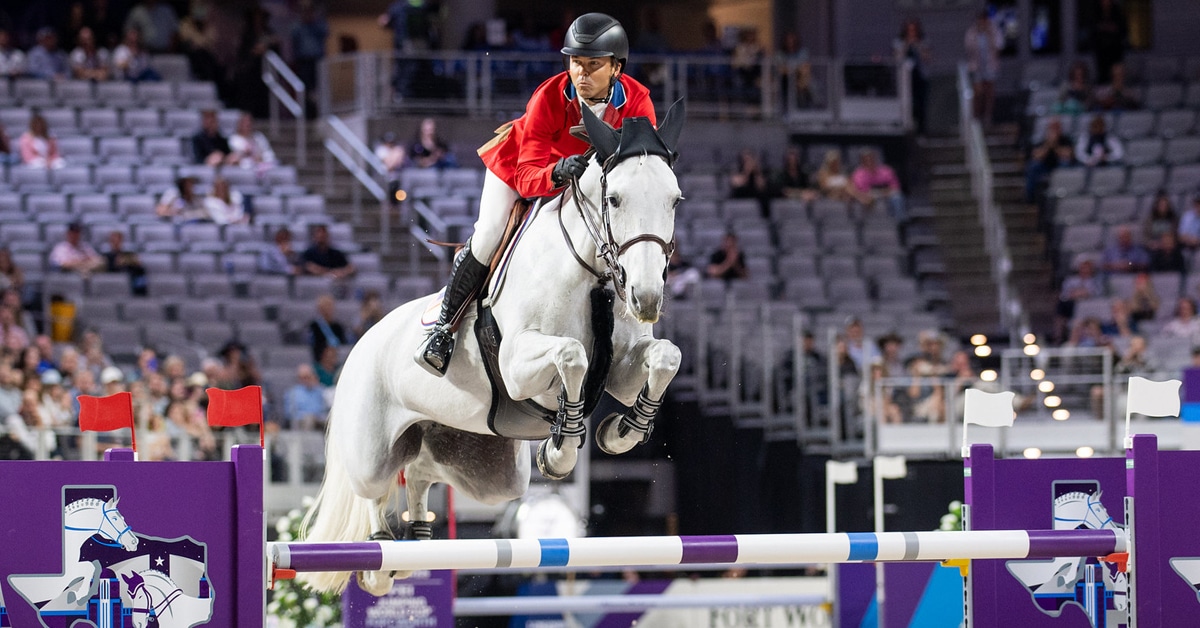 A man in a red jacket jumping a grey mare over a fence in Fort Worth.