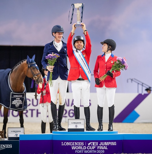 A man in a red jacket on a podium with two other riders, hoisting a large trophy.