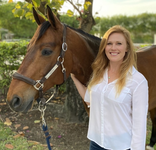 A smiling blonde woman standing with a bay horse.