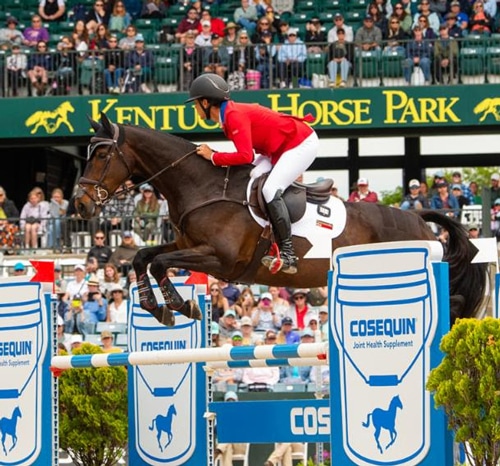 A woman in a red jacket jumping a horse over a fence in Kentucky.