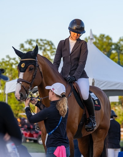 A groom adjusting the bridle of a bay horse with a female rider up.