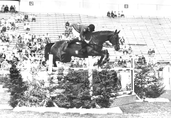 An old black-and-white photo of a man jumping a horse over a fence in a stadium.
