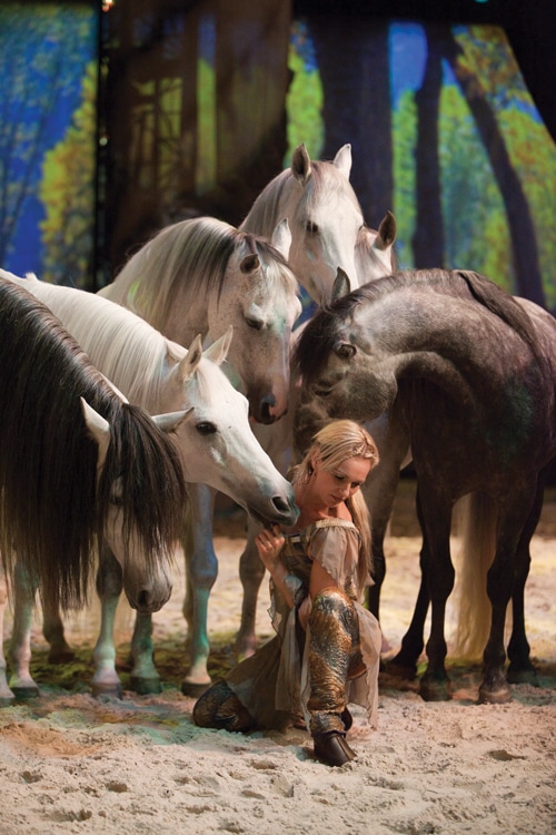 A woman crouching with white horses gathered around her.