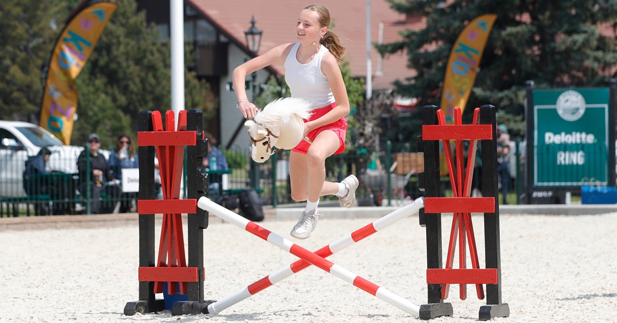 A teen girl jumping over a fence on a hobby horse.