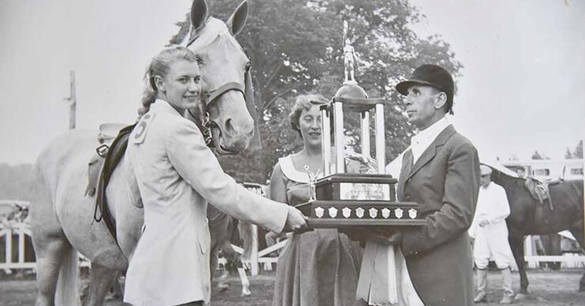 An old black-and-white photo of a woman with a grey horse being awarded a large trophy.