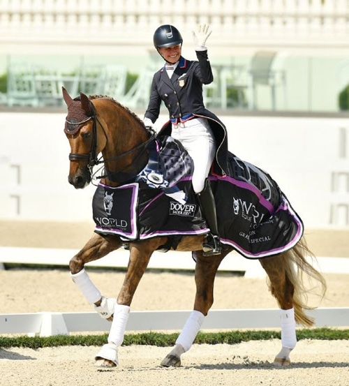 A smiling woman waving from the back of a chestnut dressage horse.