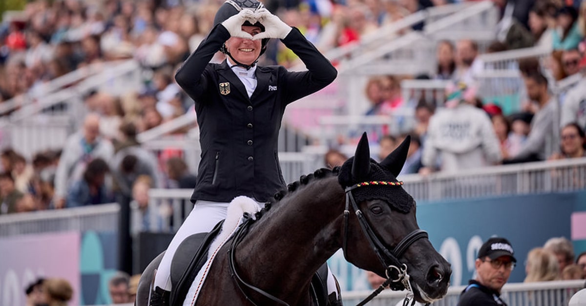 A woman on a dressage horse forming a heart with her hands.