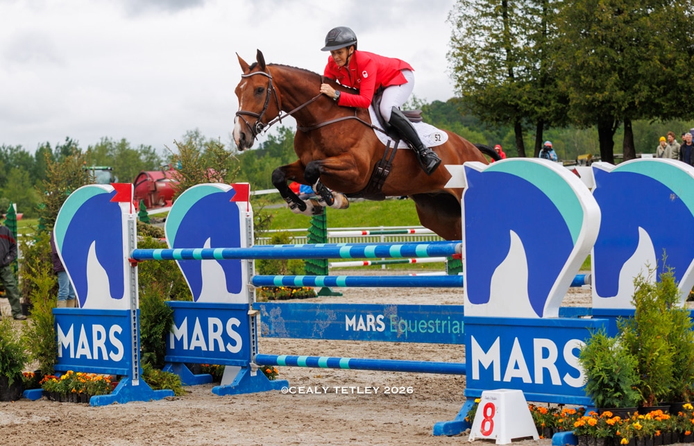 A woman in a red jacket jumping a bay horse over a fence in Bromont.