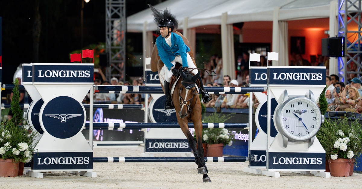 A man in a blue jacket jumping a bay horse over a fence in Cannes.