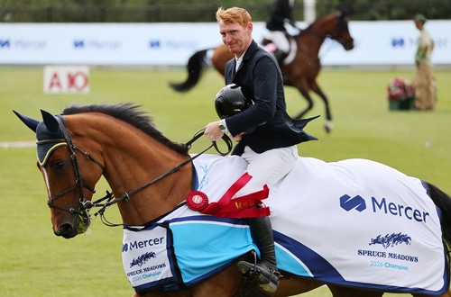 A smiling red-haired man on a bay horse during a victory gallop.