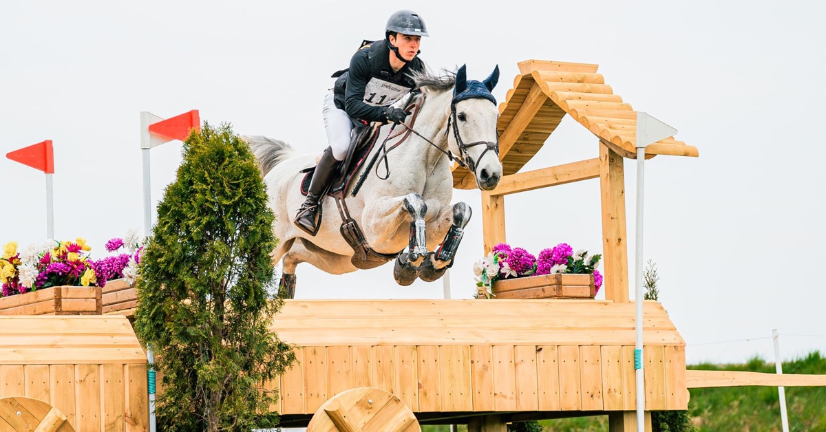 A young man jumping a grey horse over a cross-country fence.