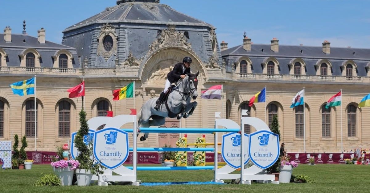 A man jumping a grey horse over a fence with a palace in the background.