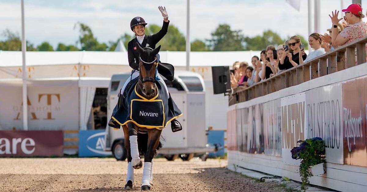 A smiling woman on a bay dressage horse, waving to the crowd.