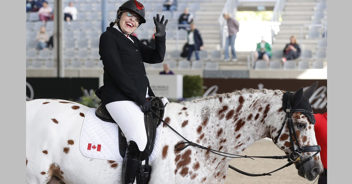 A smiling woman waving from the back of an appaloosa-coloured horse.