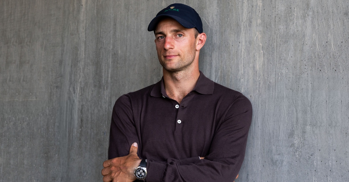 A man wearing a Rolex watch and a baseball cap posing against a grey wall.