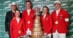 A team wearing red jackets standing with a large trophy.