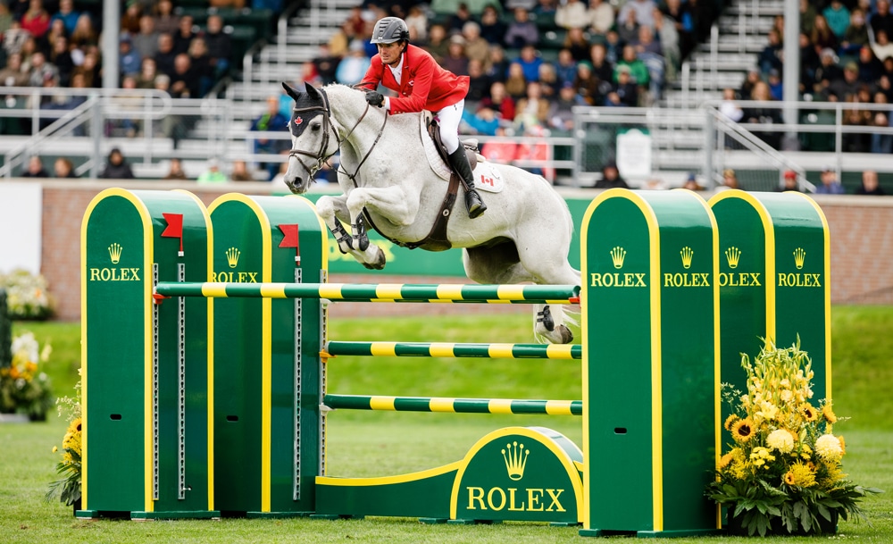 A man in a red jacket jumping a grey horse over a fence at Spruce Meadows.